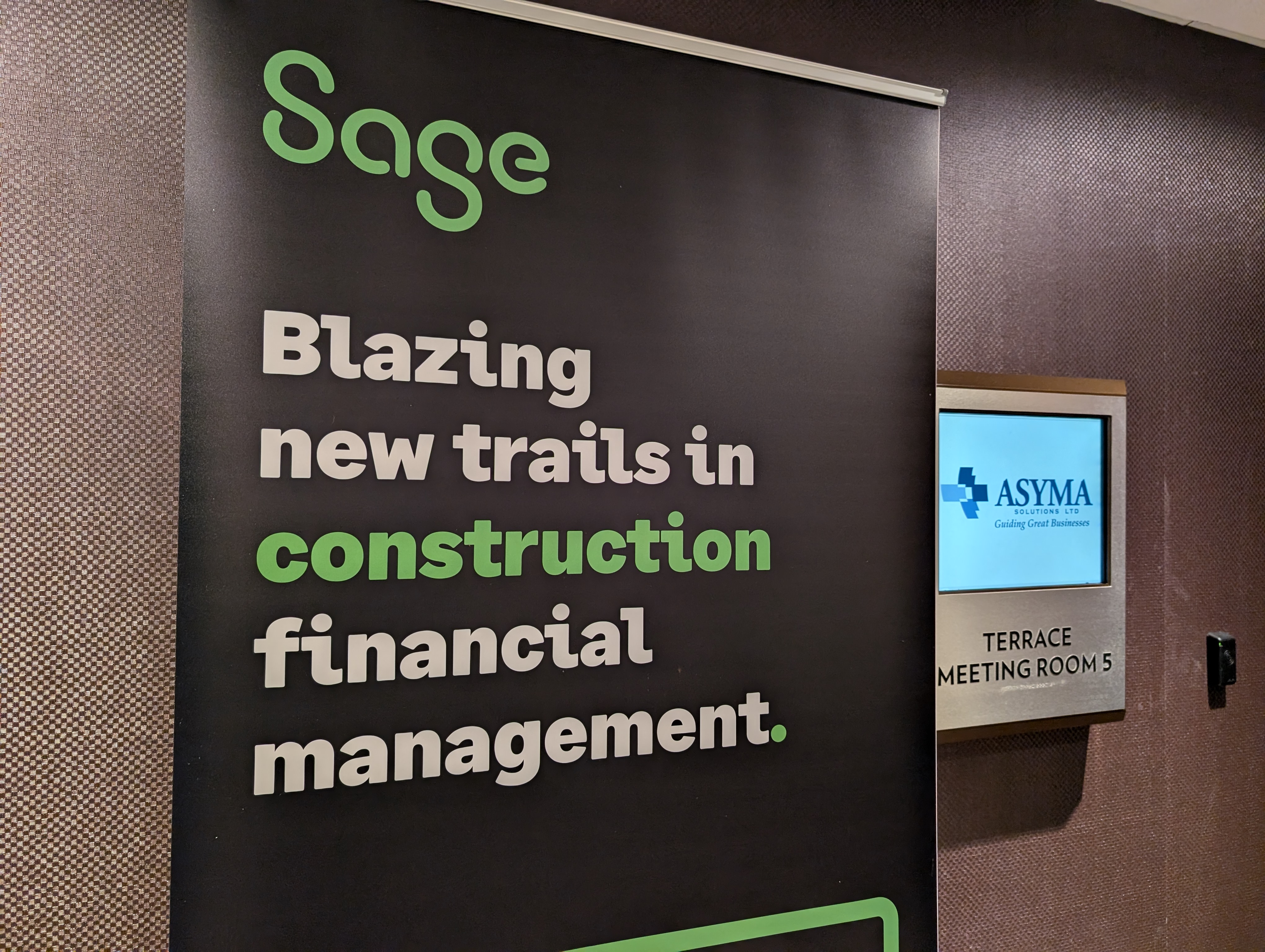Pop-up banner with Sage info on it and Asyma logo next to it on a wall display at a conference